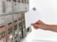 Close-up of a hand inserting a stamped envelope into a mailbox, symbolizing voting.