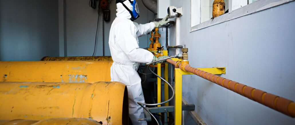 Worker in full protective suit and gas mask handles industrial pipes indoors in Indonesia.