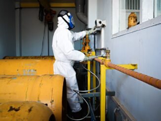 Worker in full protective suit and gas mask handles industrial pipes indoors in Indonesia.