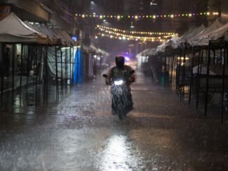 Motorcycle rider braves heavy rain in Bangkok street lined with tents and string lights at night.