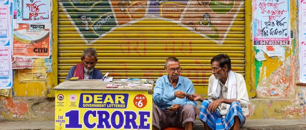 Three men sit at a lottery booth outside in Raiganj, India. Colorful posters and signs add vibrancy.