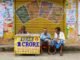 Three men sit at a lottery booth outside in Raiganj, India. Colorful posters and signs add vibrancy.
