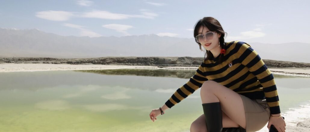 A young woman wearing sunglasses and boots poses by a scenic lake in Nanjing, China on a bright summer day.