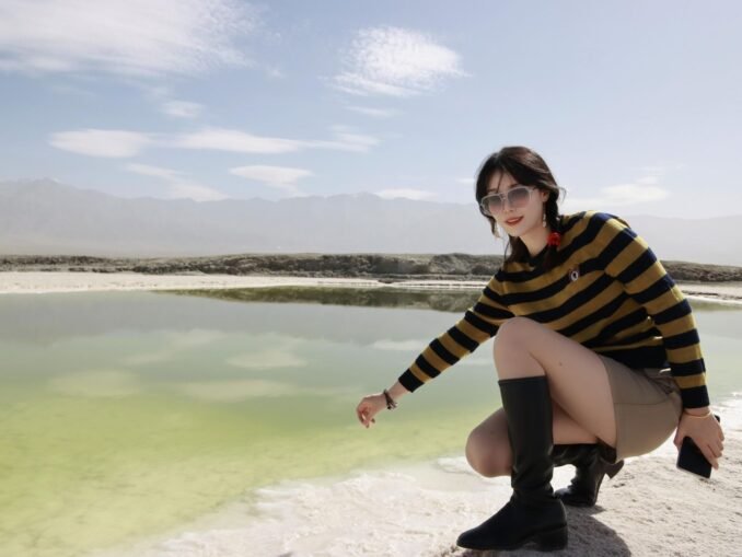 A young woman wearing sunglasses and boots poses by a scenic lake in Nanjing, China on a bright summer day.